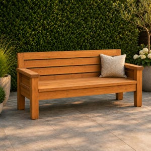 May include: A wooden outdoor bench with a light brown finish. The bench has a slatted back and armrests. A decorative pillow with a textured pattern rests on the bench. The bench is set against a backdrop of green foliage.