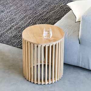 May include: A light oak side table with a circular top and vertical wooden slats. A glass of water sits on the table. The table is next to a pale blue sofa and a grey rug. The table's diameter is approximately 40 cm.