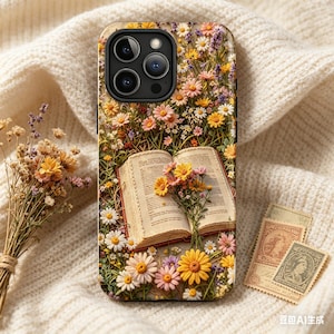 Könnte beinhalten: Eine Handyhülle mit floralem Design, die ein offenes Buch und einen Blumenstrauß zeigt. Die Hülle zeigt Gänseblümchen, Lavendel und andere Wildblumen. Der Hintergrund ist ein weicher, cremefarbener Strickstoff.