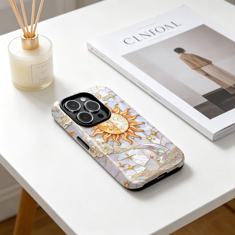 May include: A phone case with a celestial design, including a sun, moon, stars, and clouds. The case has a pearlescent finish with gold detailing. The phone case is on a white table next to a magazine and a candle.