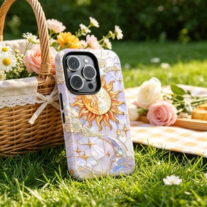 May include: A phone case with a celestial design, featuring a sun, moon, and stars. The case has a pearlescent background with gold detailing. The phone case is resting on green grass.