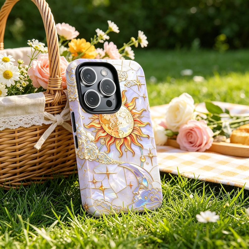 May include: A phone case with a celestial design, featuring a sun, moon, and stars. The case has a pearlescent background with gold detailing. The phone case is resting on green grass.
