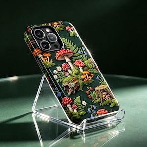 May include: A dark green smartphone case featuring a detailed forest floor pattern with red and white spotted mushrooms, small orange fungi, green ferns, tiny white daisies, blue bell-shaped flowers, wild strawberries, and a wooden tree stump.