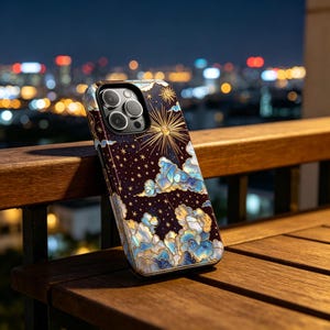 May include: A smartphone case with a celestial design. It features a dark background, gold sunburst, stars, and blue and gold clouds. The case is on a wooden surface with a city skyline in the background.