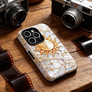 May include: A phone case with a celestial design featuring a sun, moon, stars, and clouds in gold and white. The case is on a wooden surface with film strips and cameras in the background.