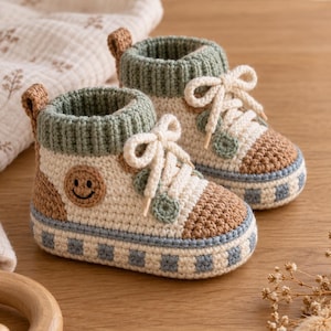 May include: A pair of crochet baby booties designed to look like high-top trainers. The booties feature a cream-coloured base, sage green ribbed cuffs, brown toe caps, and a blue and white chequered sole. A small brown smiley face patch is on the side.