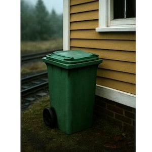 Pode incluir: Um caixote do lixo de plástico verde com uma tampa articulada e uma única roda preta. O caixote está posicionado ao lado de um edifício amarelo com uma janela. O fundo inclui trilhos de comboio e uma linha de árvores.