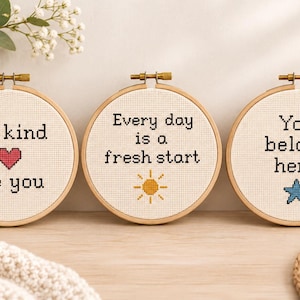 Może przedstawiać: Trzy okrągłe drewniane tamborki z białą tkaniną Aida z wyhaftowanym krzyżykiem tekstem i ikonami. Pierwszy zawiera napis Be kind Be you z czerwonym sercem. Drugi zawiera napis Every day is a fresh start ze żółtym słońcem. Trzeci zawiera napis You belong here z niebieską gwiazdą.