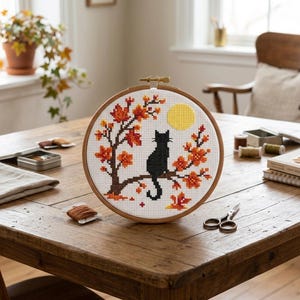 May include: A circular wooden embroidery hoop containing a cross-stitch design on white fabric. The pattern features a black cat sitting on a tree branch with orange and red autumn leaves, accompanied by a bright yellow full moon in the background.