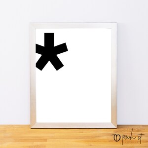 May include: A black and white minimalist art print of an asterisk in a silver frame. The print is on a white background and the frame is on a wooden surface.