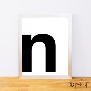 May include: A framed print featuring a large, bold, black lowercase letter "n" centered on a white background. The frame is a light wood color. The print is displayed on a wooden surface. The bottom right corner of the frame has the text "peach st design co."