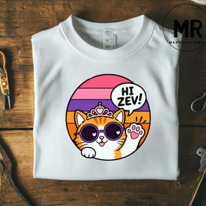 Könnte beinhalten: Weißes Sweatshirt mit einer Cartoon-Katze, die eine Sonnenbrille und eine Krone trägt, mit dem Text "HI ZEV!" in einer Sprechblase. Die Katze ist vor einem farbenfrohen, gestreiften Hintergrund aus Pink, Orange und Lila platziert.