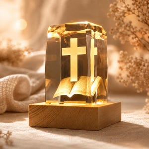 May include: A crystal block ornament with a cross and open book design, illuminated from within. The cross is a prominent feature, set on a wooden base. The faceted crystal creates a warm glow. Religious home decor item.