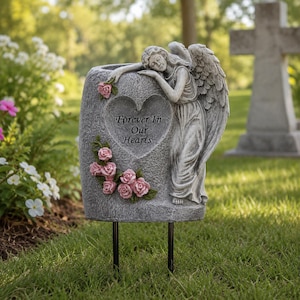 May include: A gray stone-textured memorial garden stake featuring a sculpted angel leaning against a heart-shaped plaque. The plaque is inscribed with the text Forever In Our Hearts and is decorated with several small pink roses and green leaves.