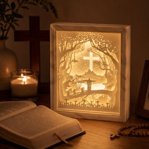May include: A rectangular light box featuring a layered paper-cut scene of a landscape with a central cross, a figure with outstretched arms, and silhouettes of trees and smaller crosses, all illuminated by a warm, soft internal glow.