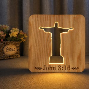 Puede incluir: Una luz de noche de madera con un recorte de la estatua del Cristo Redentor, iluminada con un brillo cálido. El texto "John 3:16" está grabado debajo. La luz es cuadrada y está hecha de madera clara.