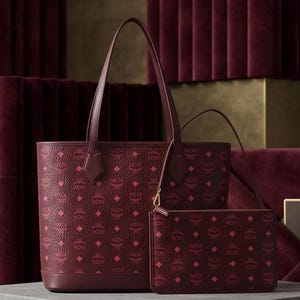 May include: Burgundy tote bag and matching clutch with a repeating pink logo pattern. The tote has long handles, and the clutch has a gold zip. The bags are displayed on a grey surface against a burgundy backdrop.