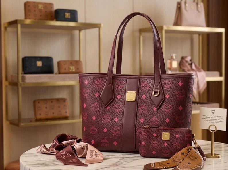 May include: Burgundy tote bag, wallet, and scarf set with a repeating pink diamond pattern. The tote has a gold-tone logo plate and matching handles. Other leather goods are displayed on shelves in the background.