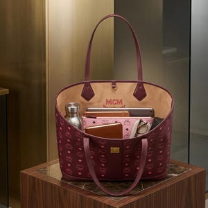 May include: Burgundy tote bag with a repeating logo pattern. The bag has a light tan interior and contains a silver water bottle, a pink wallet, a tablet, a brown wallet, and eyeglasses. The bag is on a brown wooden surface.