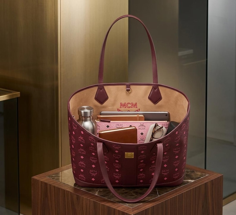 May include: Burgundy tote bag with a repeating logo pattern. The bag has a light tan interior and contains a silver water bottle, a pink wallet, a tablet, a brown wallet, and eyeglasses. The bag is on a brown wooden surface.