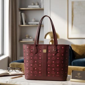 May include: Burgundy tote bag featuring a repeating pink diamond and MCM logo pattern. The bag has two burgundy handles, a vertical centre stripe, and a gold-coloured metal accent. The bag is on a table with a notebook, glasses, and a pen.