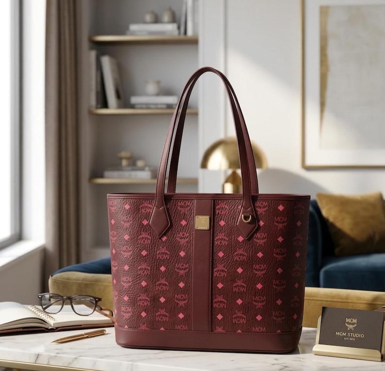 May include: Burgundy tote bag featuring a repeating pink diamond and MCM logo pattern. The bag has two burgundy handles, a vertical centre stripe, and a gold-coloured metal accent. The bag is on a table with a notebook, glasses, and a pen.