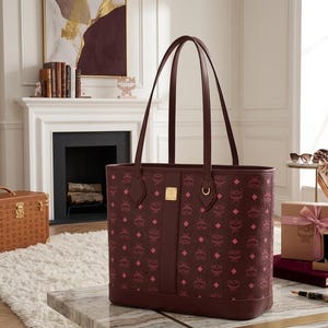 May include: A burgundy tote bag featuring a repeating pink diamond pattern and the letters "MCM". The bag has two burgundy handles and a gold metal accent. The bag is on a marble table.