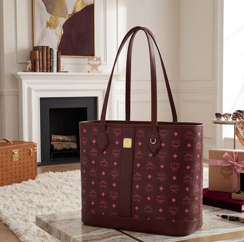 May include: A burgundy tote bag featuring a repeating pink diamond pattern and the letters "MCM". The bag has two burgundy handles and a gold metal accent. The bag is on a marble table.