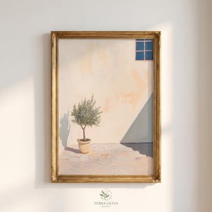 Op de afbeelding: Een ingelijste kunstdruk toont een serene buitenscène. Een kleine olijfboom in een beige pot staat op een stenen terras. Een blauw raam en een schaduw worden op de lichtgekleurde muur geworpen. De lijst is goudkleurig.