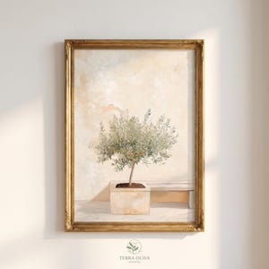 Op de afbeelding: Een ingelijste kunstdruk met een olijfboom in een vierkante pot. Het schilderij heeft een neutraal kleurenpalet en een gouden, sierlijke lijst. Het kunstwerk is onderaan gesigneerd met "Terra Oliva Prints".