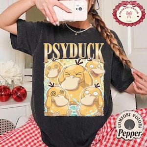 May include: A black short-sleeve t-shirt featuring the text PSYDUCK in large gold letters above a graphic illustration of five yellow Psyduck characters with various facial expressions, set against a light blue and yellow background.
