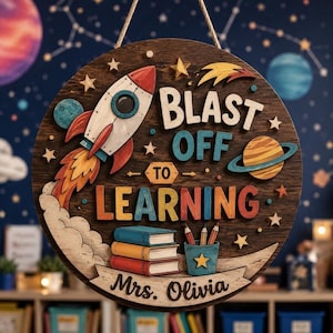 May include: A round wooden wall sign featuring a space theme with a rocket, planets, and stars. The text reads "BLAST OFF TO LEARNING" and "Mrs. Olivia". Below the text are three stacked books and a pencil holder with colored pencils.
