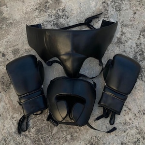 Könnte beinhalten: Ein Set schwarzer Boxausrüstung, einschließlich Handschuhen, Kopfschutz und einem Schutzbecher. Die Artikel sind aus Leder gefertigt und auf einer strukturierten, grauen Oberfläche angeordnet. Die Handschuhe haben Schnürsenkel, und der Kopfschutz bietet Ohren- und Gesichtsschutz.