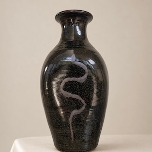 May include: A tall, bulbous ceramic vase with a glossy, dark black glaze. The surface features a single, vertical, wavy line in a muted, light grey tone that runs down the centre of the body. The vase has a narrow neck and a flared rim.