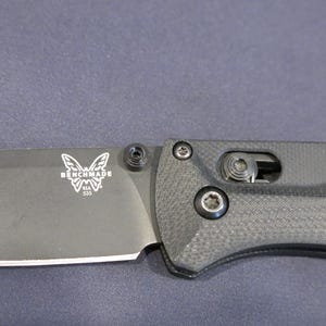 Könnte beinhalten: Ein Klappmesser mit mattschwarzer Klinge und strukturiertem schwarzem Griff. Die Klinge zeigt ein weißes Schmetterlingslogo mit dem Text BENCHMADE U.S.A. 535. Der Griff enthält eine sichtbare Pivot-Schraube und einen Metall-Verriegelungsmechanismus.