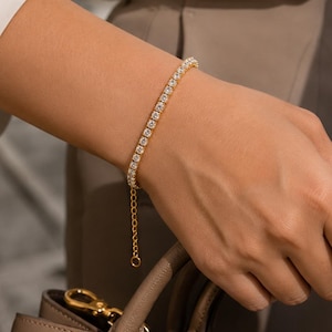 May include: A gold-toned tennis bracelet featuring a continuous row of clear, round-cut stones set in a metallic frame. The bracelet includes an adjustable chain extension with a small circular link at the end, resting on a wrist above a handbag.