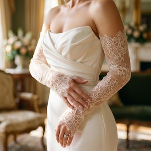 Könnte beinhalten: Ein trägerloses elfenbeinfarbenes Kleid mit einem figurbetonten Oberteil mit diagonalen Falten und langen, transparenten Spitzenärmeln, die bis zu den Fingern reichen. Die Ärmel bestehen aus zarter Blumenspitze mit gewellten Kanten an den Handgelenken.