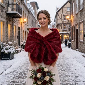 May include: A deep red faux fur stole worn over a white dress, featuring a thick, plush texture that wraps around the shoulders. The stole is paired with a floral bouquet containing red and white roses, green foliage, and blue thistle accents.
