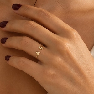 May include: A gold-toned open ring featuring two capital letters, A and E, positioned at each end of the band. The ring has a thin, polished metal construction that wraps partially around the finger, showcasing the distinct letter design clearly.