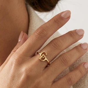 May include: A gold-toned ring featuring two interlocking letters, specifically a stylized C and D, forming a continuous band design. The metal has a polished finish, and the ring is worn on the ring finger, showcasing the metallic luster and design.