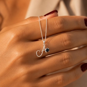 May include: A silver-toned necklace featuring a cursive letter A pendant. The pendant is accented with a single, small, round, dark blue gemstone set into the side of the letter. The necklace hangs from a delicate, thin silver-toned chain.