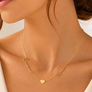 May include: A gold-toned necklace featuring a delicate chain with two script-style names, Caroline and Amelia, integrated into the design, accented by a small, centered gold-toned heart charm suspended between the two names.