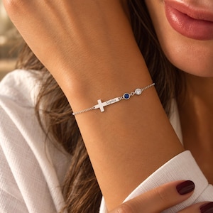 May include: A silver-toned chain bracelet featuring a horizontal cross charm engraved with the name Sophia, followed by a small dark blue circular stone and a clear white circular stone set in silver-toned metal links on a delicate chain.