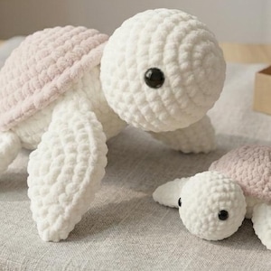 May include: Two handmade crochet sea turtles featuring soft white yarn bodies and light pink shells. Each turtle has round black plastic eyes. The larger turtle is positioned on the left, while the smaller turtle is placed to the right.