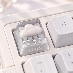 May include: A custom mechanical keyboard keycap featuring a clear resin housing with an embedded white cloud and four dangling crystal rain drops. The word Una is printed in white at the base of the keycap, which is set among other white keys.