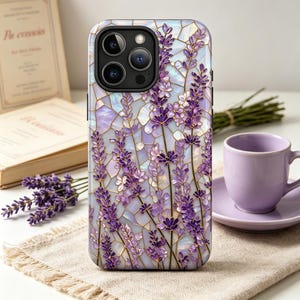 Könnte beinhalten: Eine Handyhülle mit einem Lavendel-Blumenmuster. Die Hülle hat einen mosaikartigen Hintergrund mit lila und weißen Blumen. Die Handyhülle liegt auf einer beigen Oberfläche mit einer Tasse und Untertasse im Hintergrund.