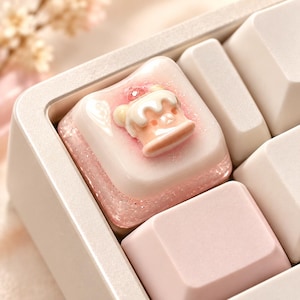 Puede incluir: Una tecla de teclado mecánico cuadrada con una base rosa claro brillante con purpurina incrustada. Centrada en la superficie superior hay una pequeña figura esculpida en tres dimensiones de un pastel con forma de oso.