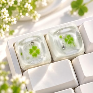 May include: Two clear, square resin keycaps installed on a white mechanical keyboard. Each keycap encapsulates a bright green four-leaf clover and a small, dangling silver-toned charm, creating a suspended, layered effect within the transparent material.