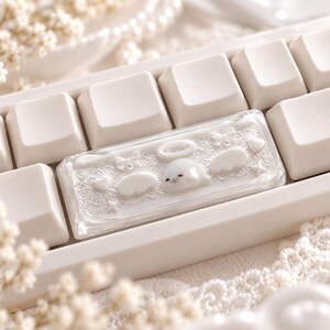 以下が含まれることがあります： 透明な樹脂仕上げの長方形の白いメカニカルキーボード用スペースバーキーキャップ。中央には天使の輪をつけた白いアザラシのフィギュアがあり、その周囲には白いレース模様、小さなリボン、ハート、天使の羽が立体的にあしらわれています。