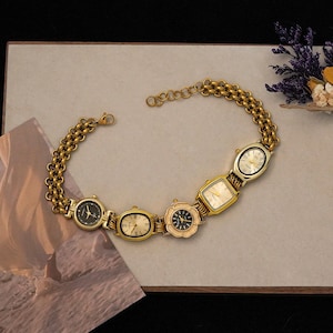 May include: A gold-coloured necklace with several vintage watch faces of different shapes. The watch faces have various dial colours, including black, cream, and gold. The necklace is displayed on a neutral surface with dried flowers in the background.